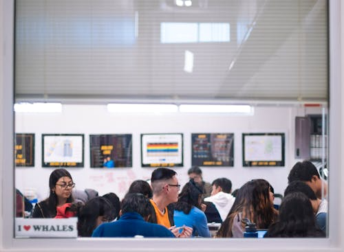 Students sit in a classroom with framed charts on the walls. A window shows the message "I ♥ WHALES." The mood is focused and studious.