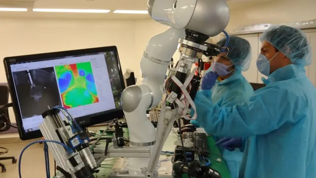 Two lab technicians in blue gowns and masks operate a robotic arm beside a monitor displaying colorful data in a high-tech lab.