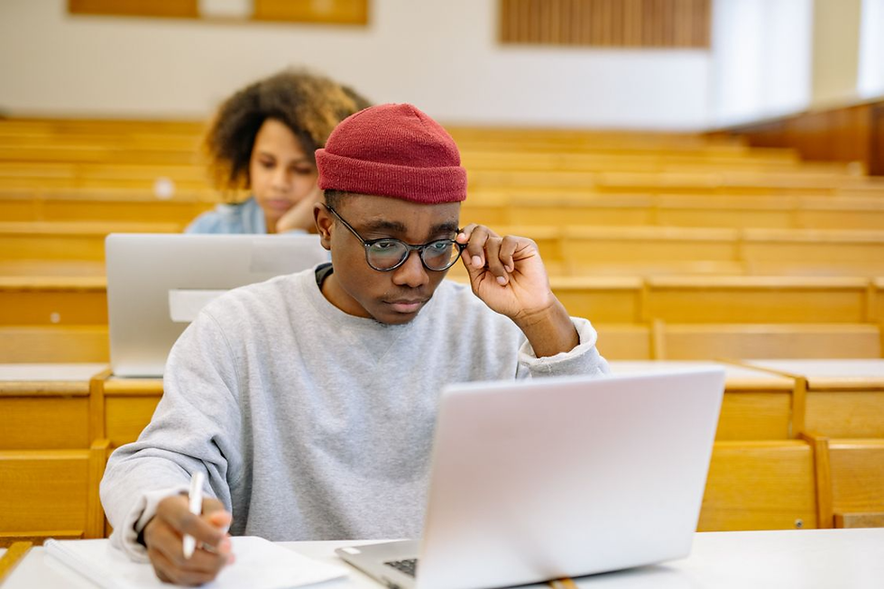 Student in red beanie studies on a laptop in a lecture hall, writing notes while another student works behind him.