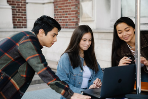 Three students smile and work together around a laptop outdoors beside a brick building.