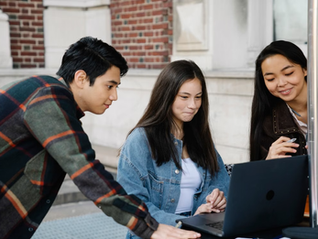 Three students smile and work together around a laptop outdoors beside a brick building.