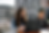 Two students sit outdoors at a table, focused on a laptop, with urban buildings blurred in the sunny background.