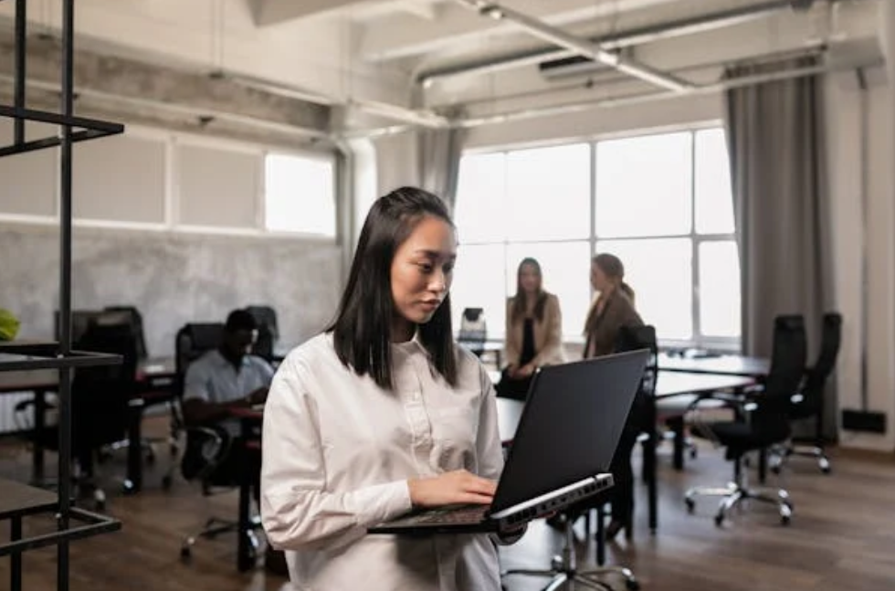 Woman using laptop in modern office with large windows. Two people in conversation in the background. Bright and professional setting.