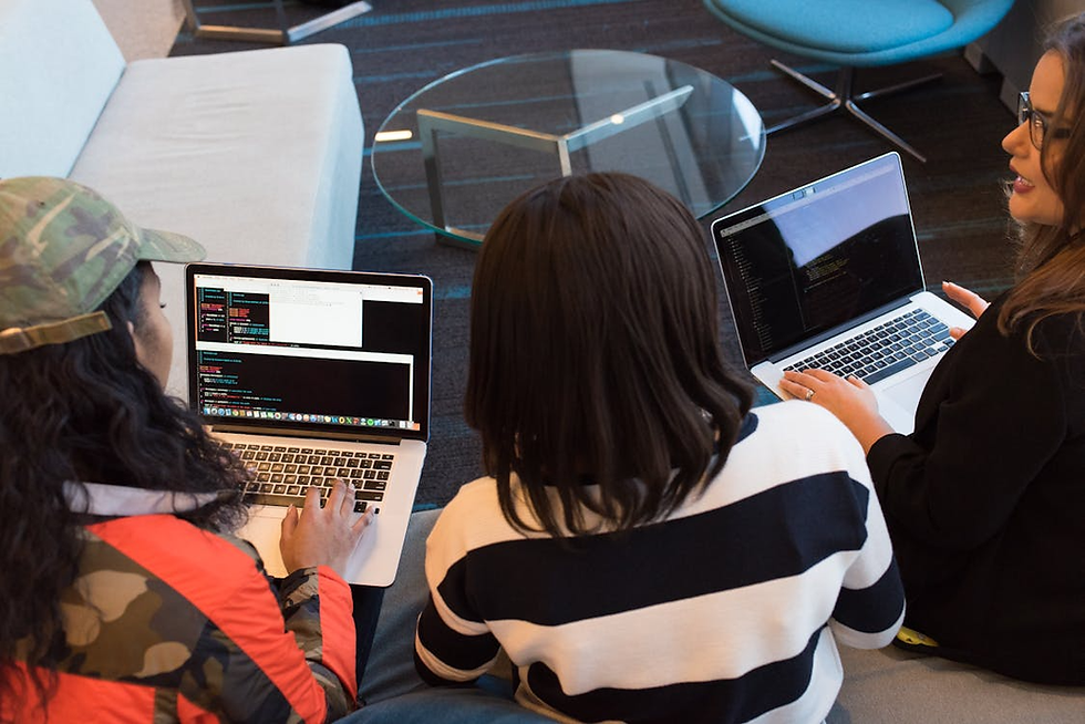 Three people sitting on a couch, coding on laptops in a modern office. One wears camouflage, another a striped sweater. A glass table nearby.