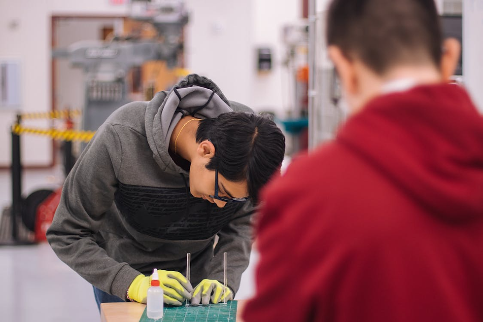 Two people in a workshop; one in focus wearing gloves, cutting material on a green mat, another blurred in red. Tools and machines in background.