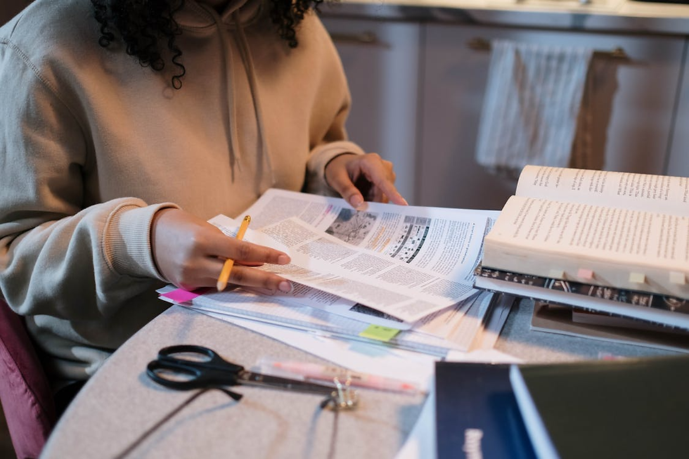 Person in a beige hoodie studying at a table with papers, books, and scissors. They hold a pencil, surrounded by a cozy kitchen setting.