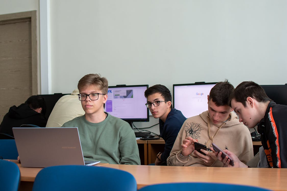 Four male students in a computer lab working on laptops and smartphones with desktop monitors in the background.
