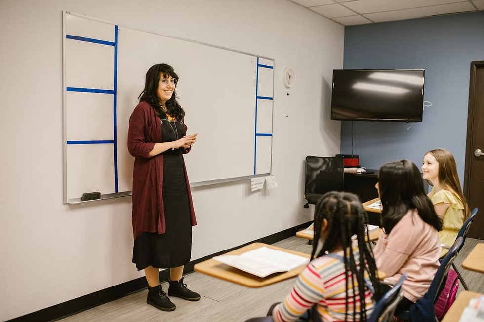 Teacher smiles beside a whiteboard while students sit at desks in a classroom with a TV on the wall.