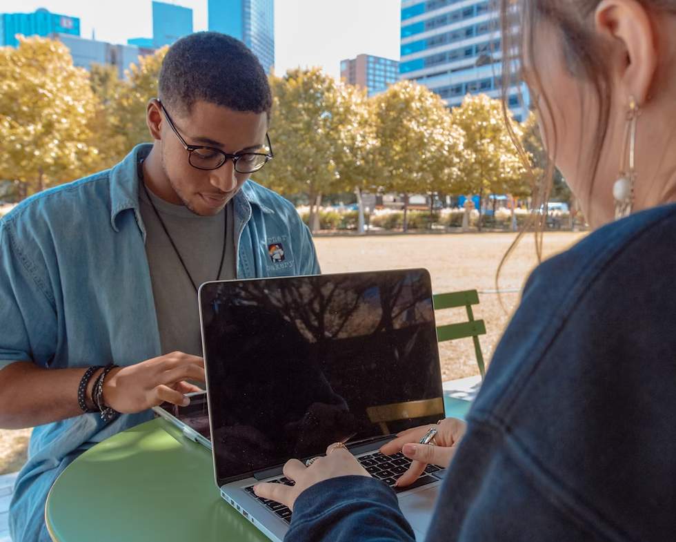 Two people work on laptops at a green table in a park, with trees and city buildings in the background under a clear sky.