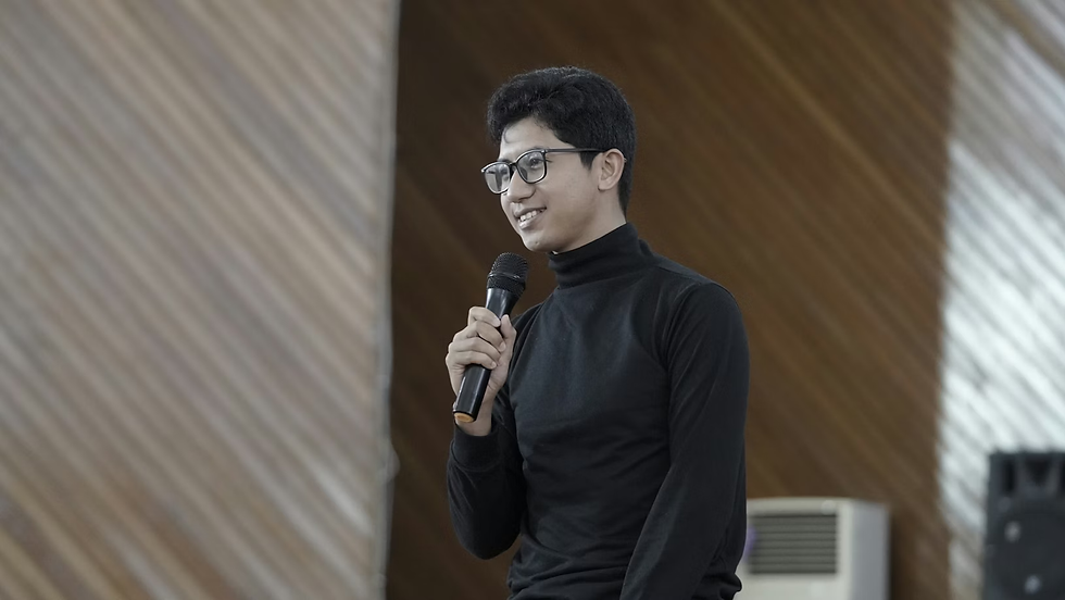 A person in a black turtleneck speaks into a microphone, smiling. Background features diagonal wooden patterns and an air conditioner.