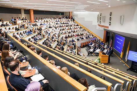 Audience in a large lecture hall listens attentively to a speaker at a podium. Presentation slides are visible on a screen.