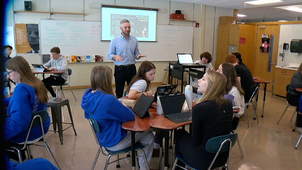 A teacher addresses students seated at desks with laptops in a classroom. A projected screen and whiteboard with notes are in the background.