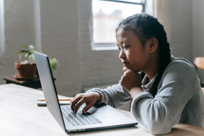 Young girl types on a laptop at a table, chin in hand, in a bright room with a window and potted plants.