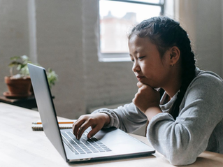 Young girl types on a laptop at a table, chin in hand, in a bright room with a window and potted plants.
