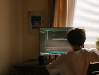 Boy seen from behind coding on a desktop monitor in a warm, dim room by a bright window and potted plant