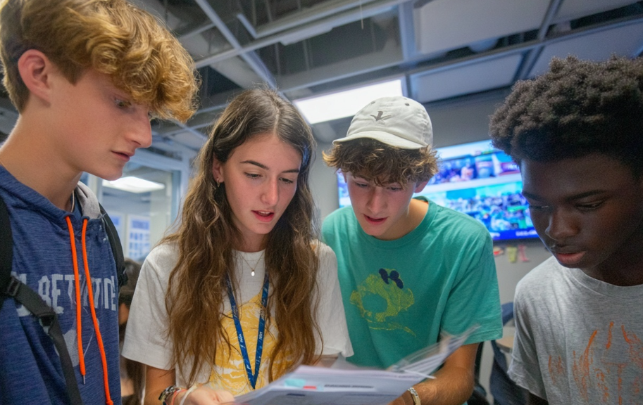 Four students closely read documents in a classroom, focused and engaged. A ceiling with lights, screens, and a window are in the background.