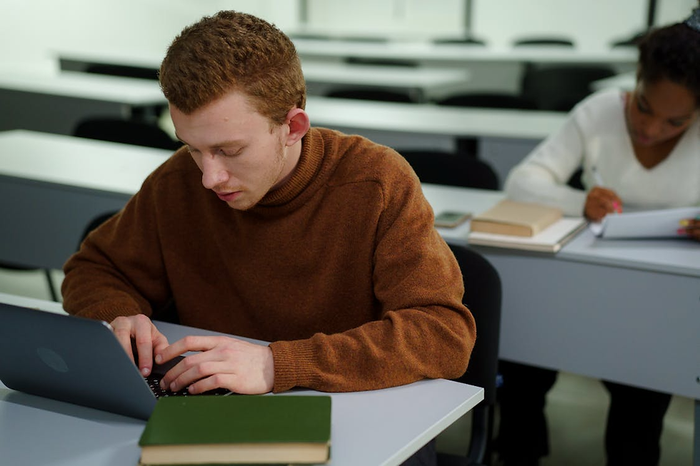 Students in a classroom focused on their tasks; one types on a laptop, another writes notes. The setting is bright with a studious mood.