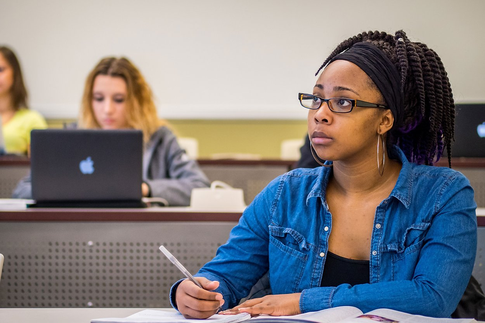 Student in a denim shirt attentively takes notes in a classroom. Other students with laptops are blurred in the background. The mood is focused.