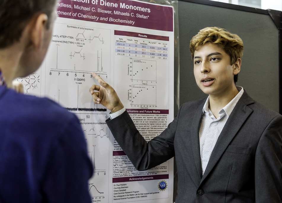 Young man in suit explains scientific poster with graphs to another person in blue. The setting is a conference; mood is focused and engaged.