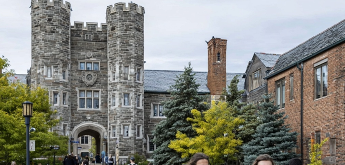 Stone building with towers in a university setting, surrounded by trees. Several people walk in the courtyard. Overcast sky, historic feel.