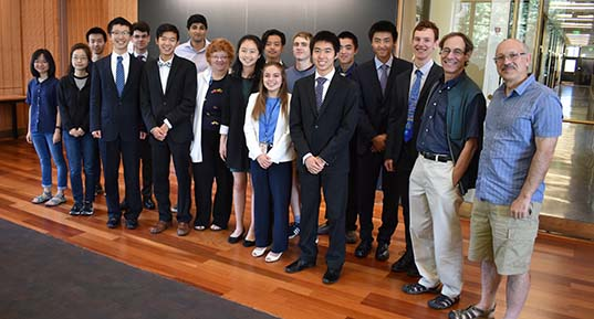 A group of people, mostly in formal attire, stand smiling on a wooden floor in front of a chalkboard. Bright, natural light fills the room.