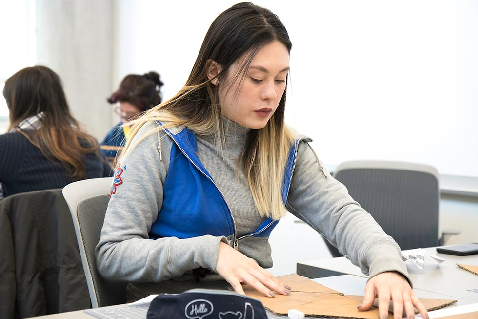 Woman in a blue jacket focuses on cutting cardboard in a classroom. Others work in the background, creating a focused, studious mood.