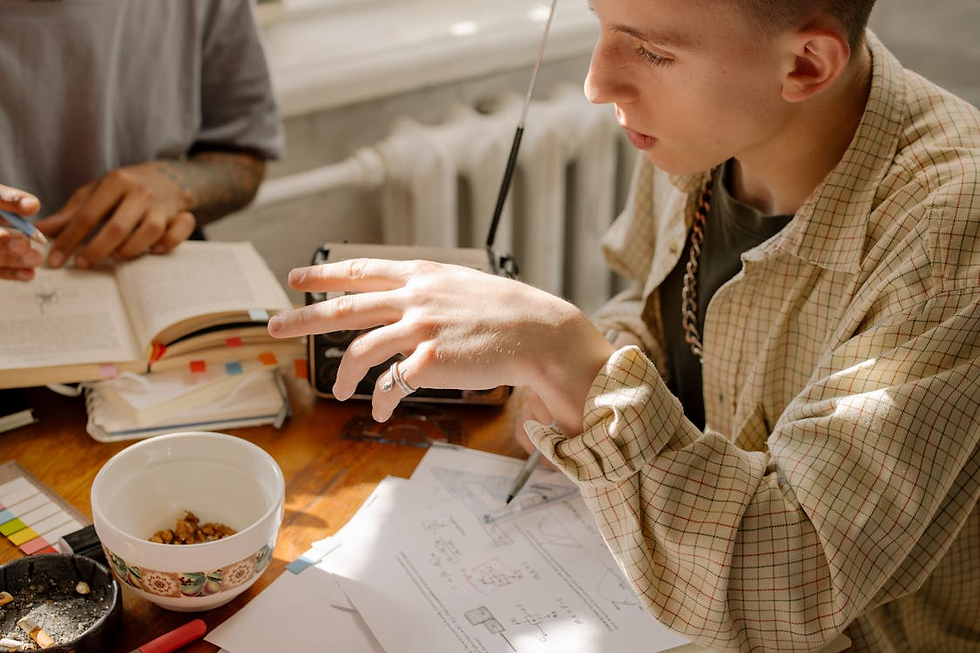 A person in a checkered shirt gestures over papers with diagrams on a sunlit wooden table. Notes, books, a bowl, and a radio are present.