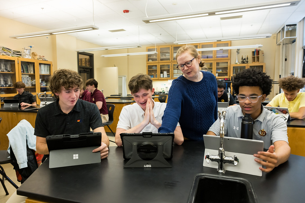 Teacher helping three students with tablets in a classroom. One student looks delighted. Background shows shelves with books and equipment.
