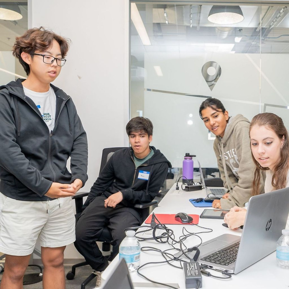 Four people in a modern office, engaged around a laptop. One stands, others sit. Whiteboard, purple water bottle, cables visible. Focused mood.