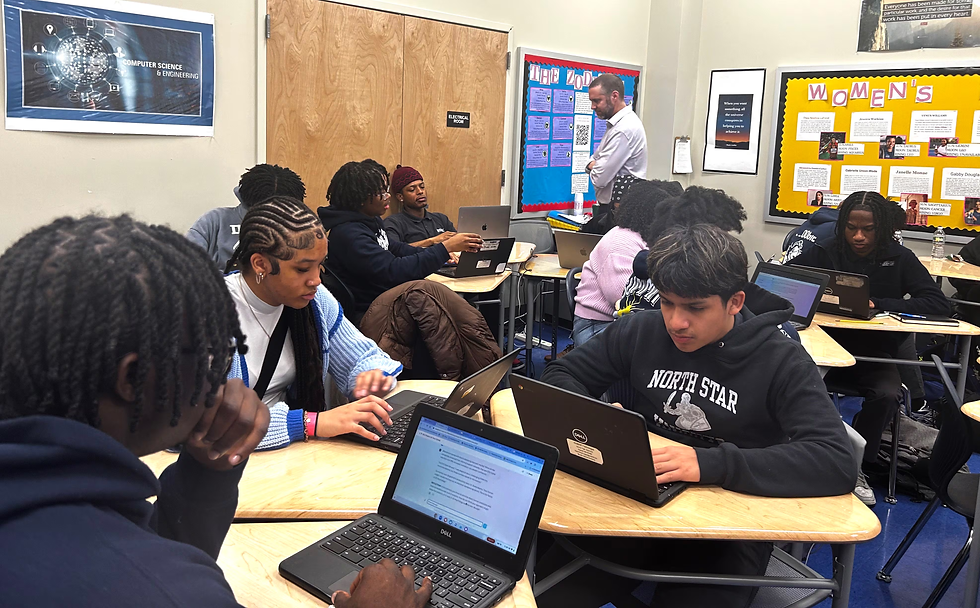 Students in a classroom work on laptops with a teacher nearby. Posters decorate the walls. Focused atmosphere, casual attire.