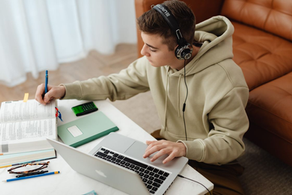 Teen boy in headphones studies at a table, writing in a book beside a laptop, notebook, calculator, and glasses on a couch.