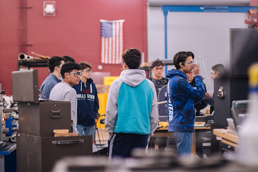 Students wearing goggles stand in a workshop with tools. An American flag hangs on a red wall. Casual attire and focused mood.