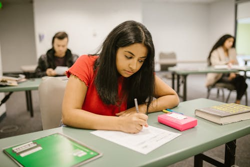 Student in a red shirt writes on paper in a classroom, seated at a green desk. Books and stationery are visible. Focused atmosphere.