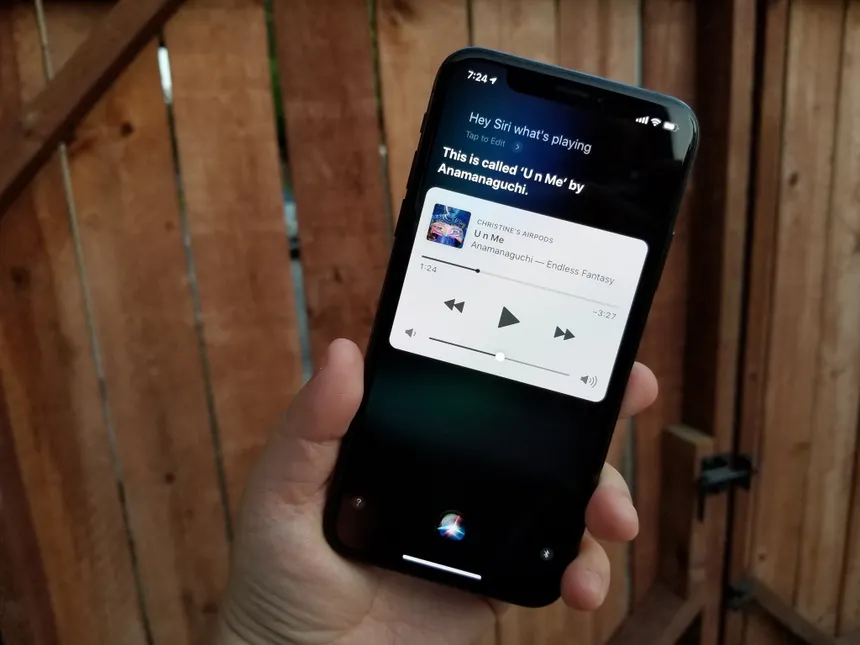 Hand holding a smartphone displaying Siri playing "U n Me" by Anamanaguchi. Wooden fence background, dim lighting, casual setting.