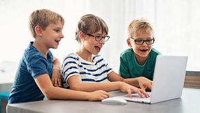 Three children excitedly look at a laptop screen. They are indoors, with one child wearing a striped shirt and the others in blue and green.
