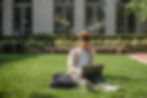 Person sitting on grass with a laptop, wearing a striped shirt. Backpack nearby. Background features bushes and building windows.