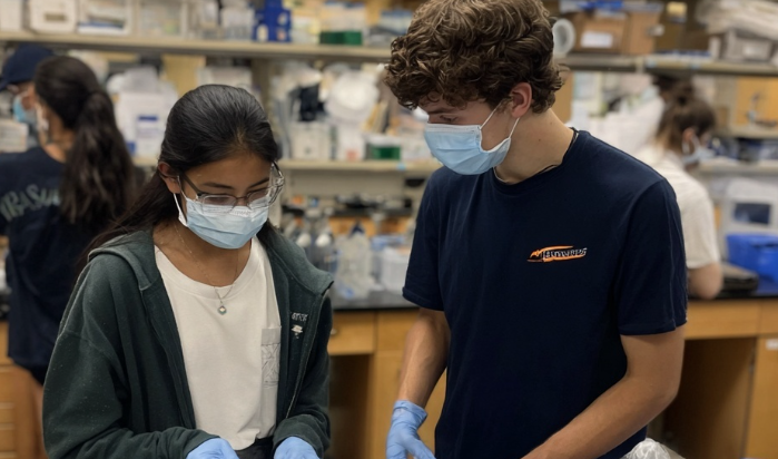 Masked students in a science lab look down at a shared task, wearing gloves amid shelves of supplies.