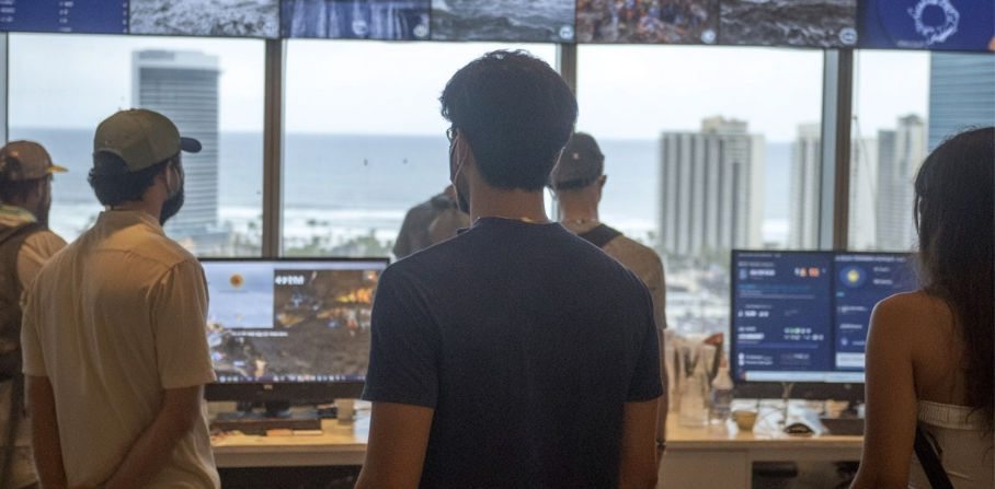People stand in an office with large windows, viewing screens showing data. Buildings and the ocean are visible in the background.