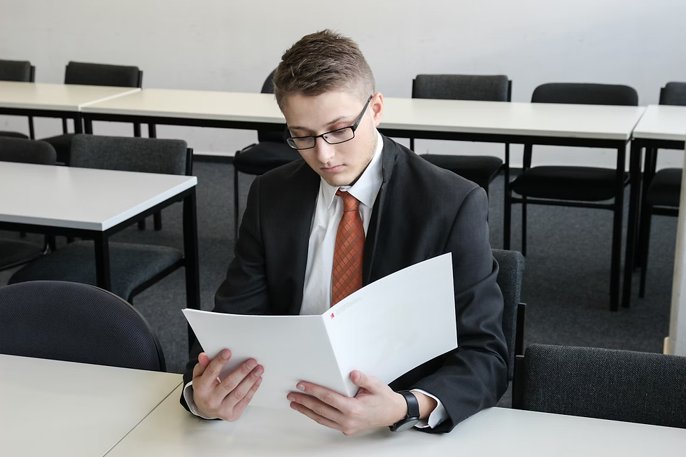 Young man in a suit reads a document in an empty classroom. He appears focused. The room has white walls, gray chairs, and tables.