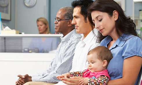 Multiple patients waiting at a healthcare facility in a waiting room at a hospital. Mother holding a baby in her lap in the forefront.