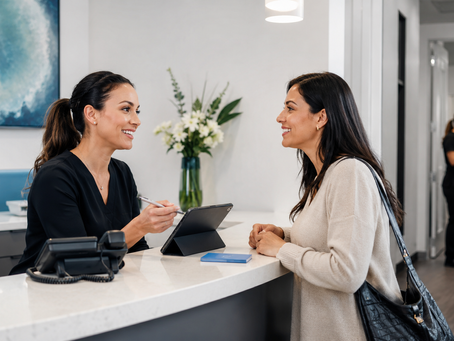 Smiling receptionist talks with a woman at a modern clinic desk, with a tablet, phone, flowers, and a staffer in back.