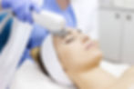Woman receiving a facial treatment with a laser device. She wears a white headband, lying on a pillow. Technician in blue gloves. Peaceful setting.