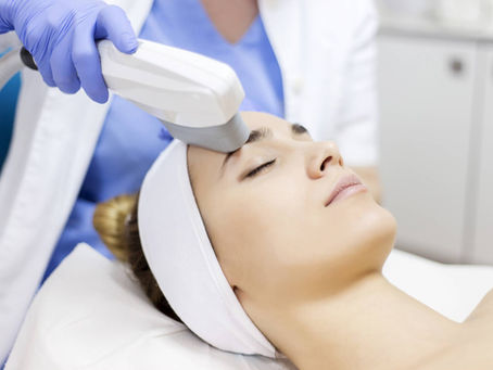 Woman receiving a facial treatment with a laser device. She wears a white headband, lying on a pillow. Technician in blue gloves. Peaceful setting.