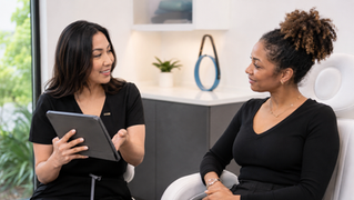 Two women in black outfits smile and talk in a bright office, one holding a tablet, with a plant and decor in the background