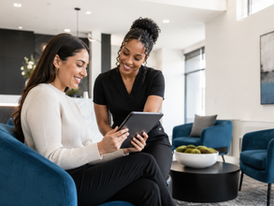 Two women smile over a tablet in a modern lobby with blue chairs, a marble desk, and abstract wall art.