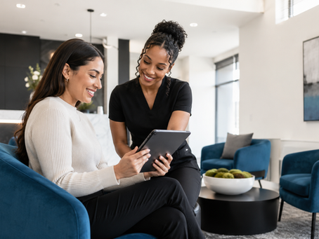 Two women smile over a tablet in a modern lobby with blue chairs, a marble desk, and abstract wall art.