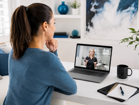 Woman in blue sweater watches a medical aesthetic virtual event on a laptop in a bright home office, with notebook, mug, and plants nearby.
