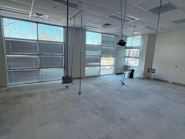 Unfinished commercial office space with exposed concrete floors, hanging electrical wiring, and large windows with blinds, shown mid-demolition prior to buildout.