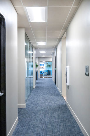 Updated office corridor with blue patterned carpet, bright white walls, modern ceiling lighting, glass partitions, and a welcoming view into a lounge area at the end of the hall.