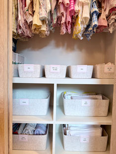Inside the closet of a luxury nursery with baby girl clothes hanging from the rod with felt baskets on the shelves above and below with pink labels.